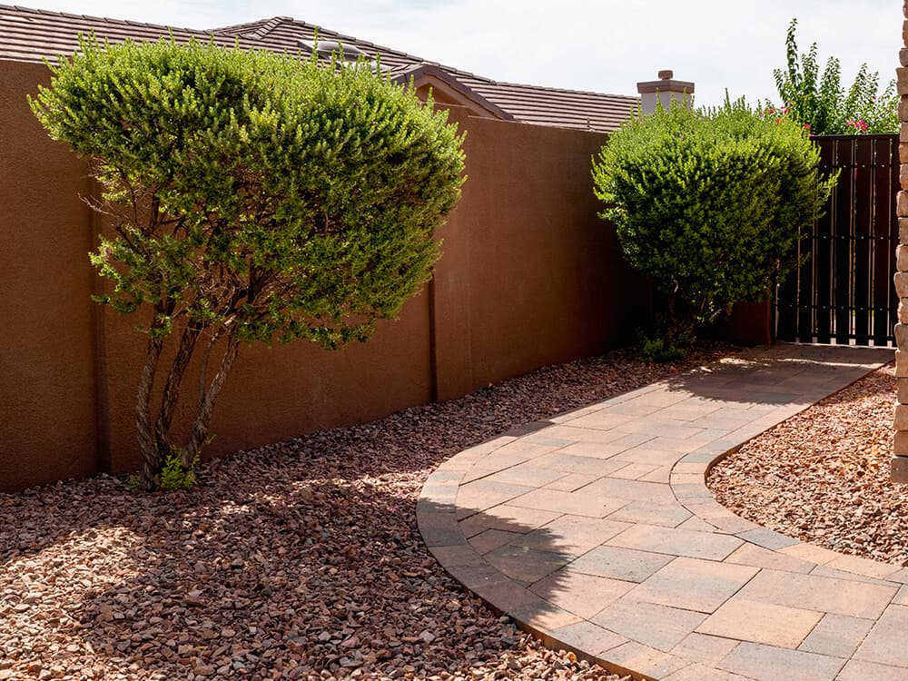 Curved paver walkway winding between shrubs and a bark bed beside a stucco wall.